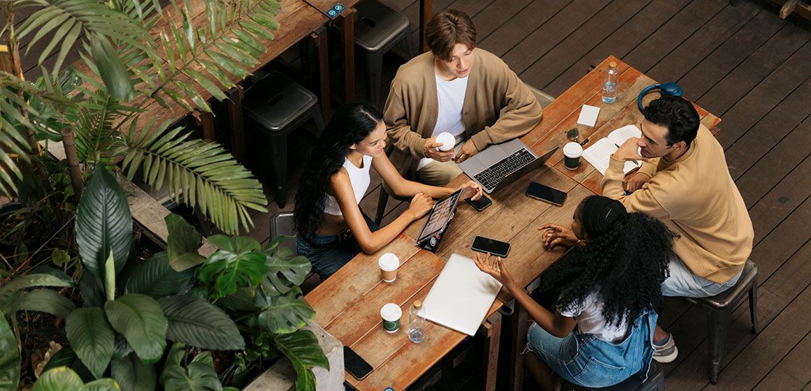 Fire kollegaer samarbeider rundt et trebord med laptoper, nettbrett og kaffe, omgitt av planter.