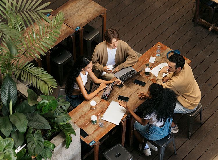Fire kollegaer samarbeider rundt et trebord med laptoper, nettbrett og kaffe, omgitt av planter.