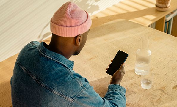 en mann med rosa lue og blå skjorte sitter på ved et bord og ser på mobilen sin