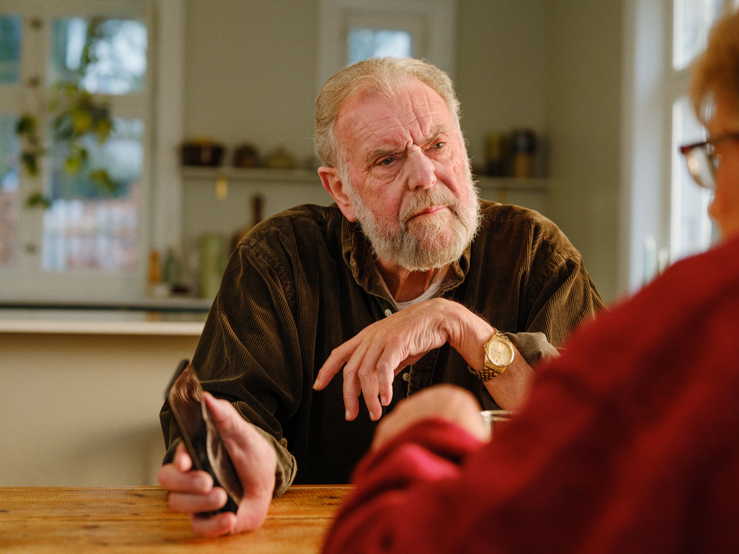 En eldre mann som sitter ved kjøkkenbordet og viser noen noe på telefonen. 