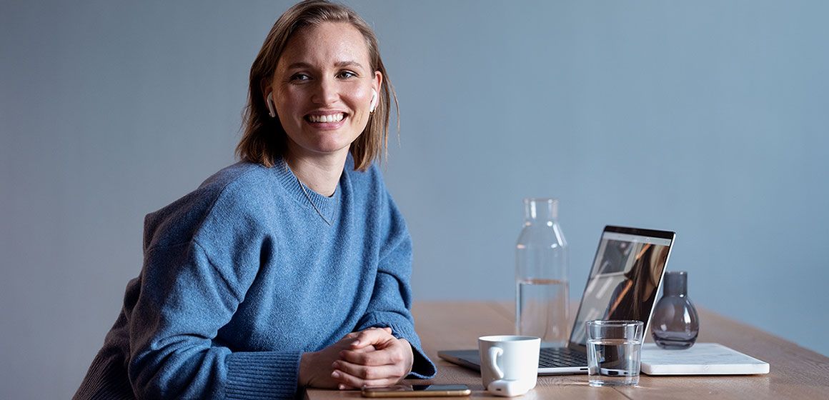 Smilende kvinne med ørepropper sitter ved et skrivebord med bærbar PC, mobil og kaffekopp.