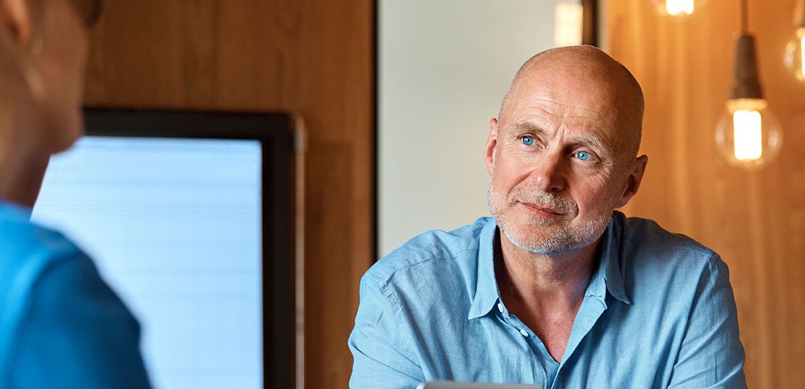 En skallet mann i lyseblå skjorte lytter oppmerksomt til en person som sitter overfor ham.