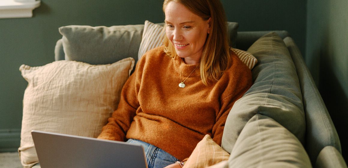 Kvinne i oransje genser sitter på sofaen med en laptop