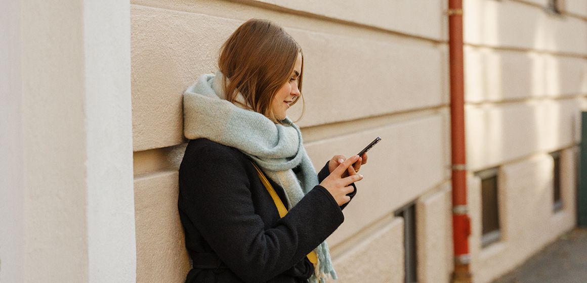 en jente som står inntil en vegg på gaten og ser på mobilen sin