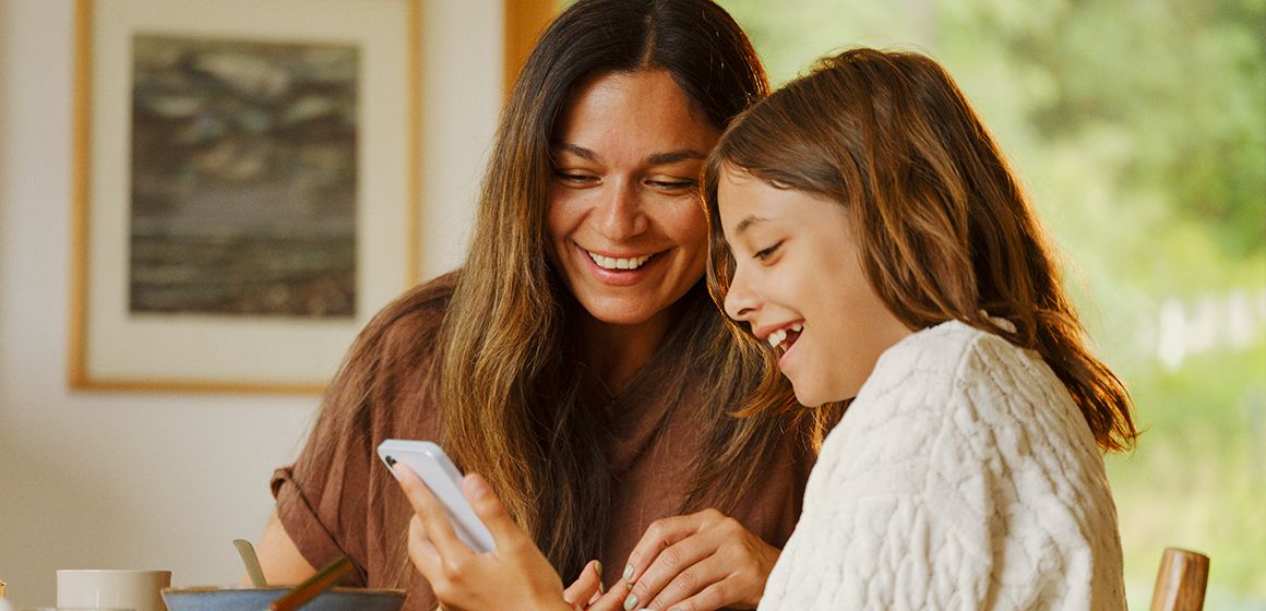Mor og datter sitter ved frokostbordet og ser på noe morsomt på mobilen