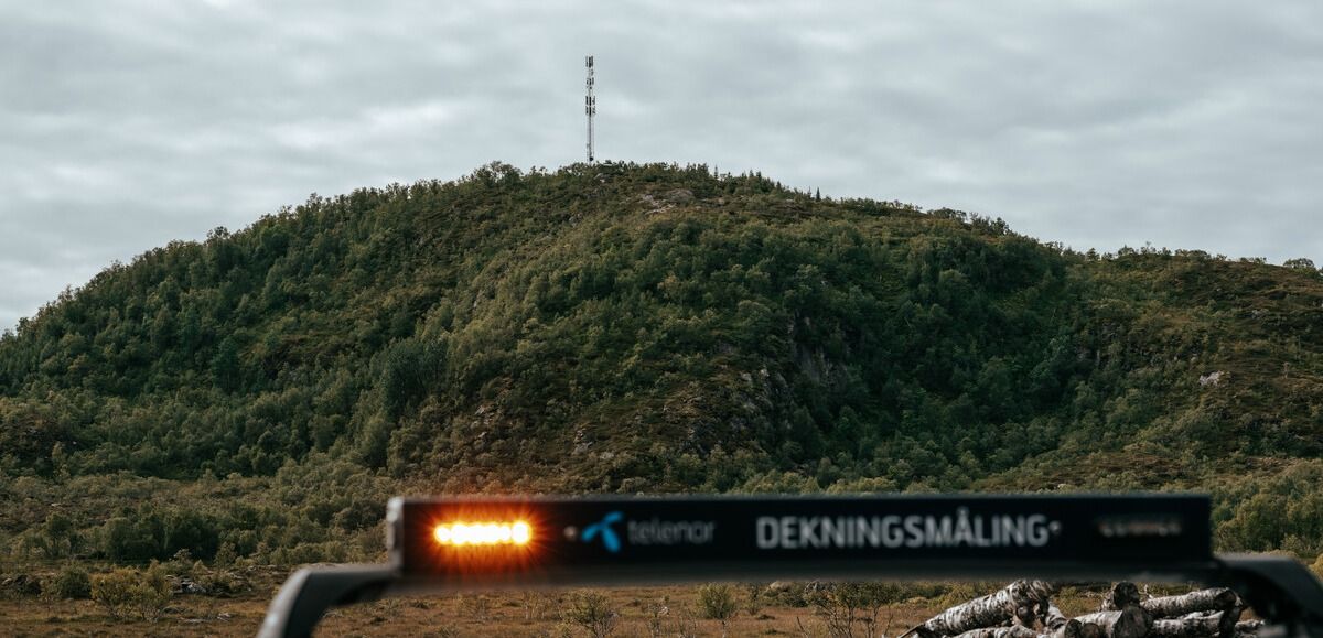 Bil med Telenor-dekningsmåling foran skogkledd ås med mastetop