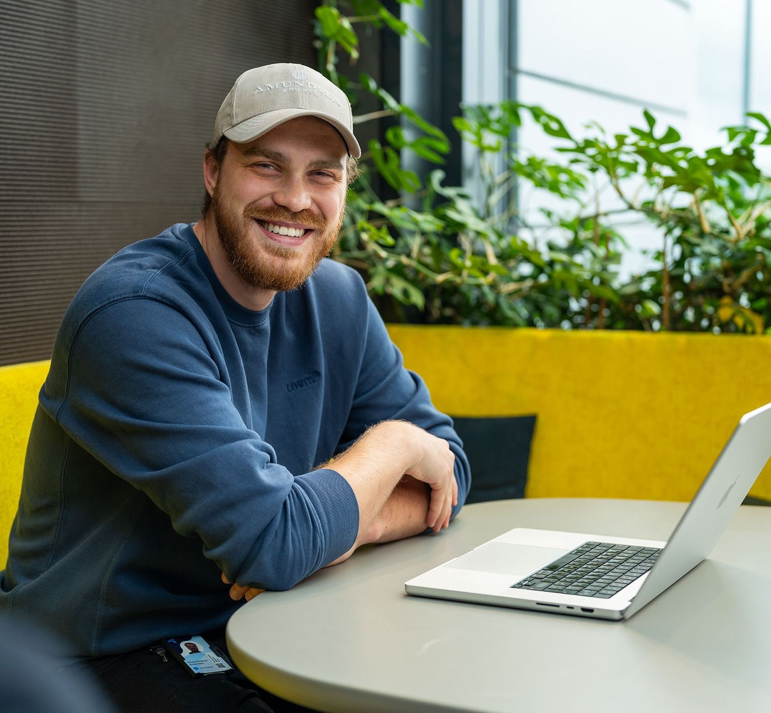 Person i blå genser ved et bord med en laptop, omgitt av planter og gult sete