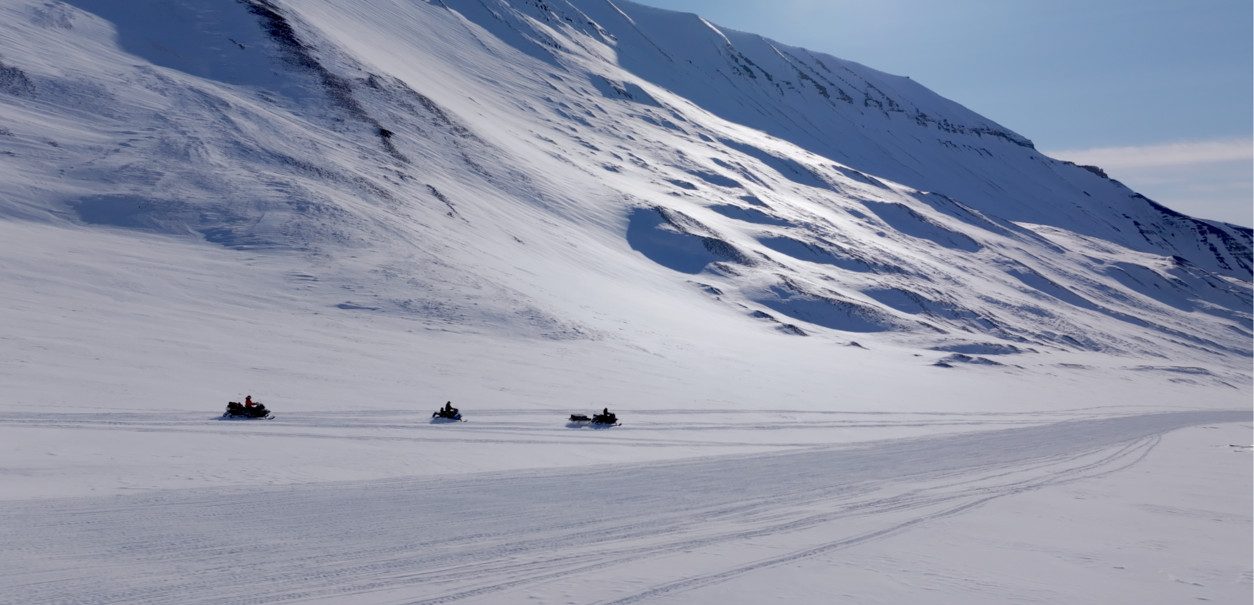 Snøscootere kjører over et snødekt landskap med fjell i bakgrunnen