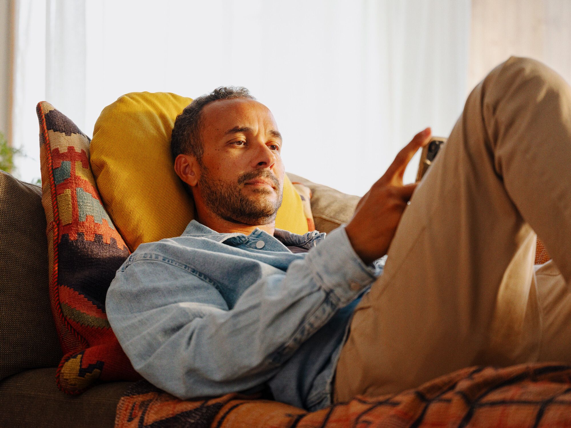 Person som ligger på en sofa med fargerike puter og pledd, holder en mobil