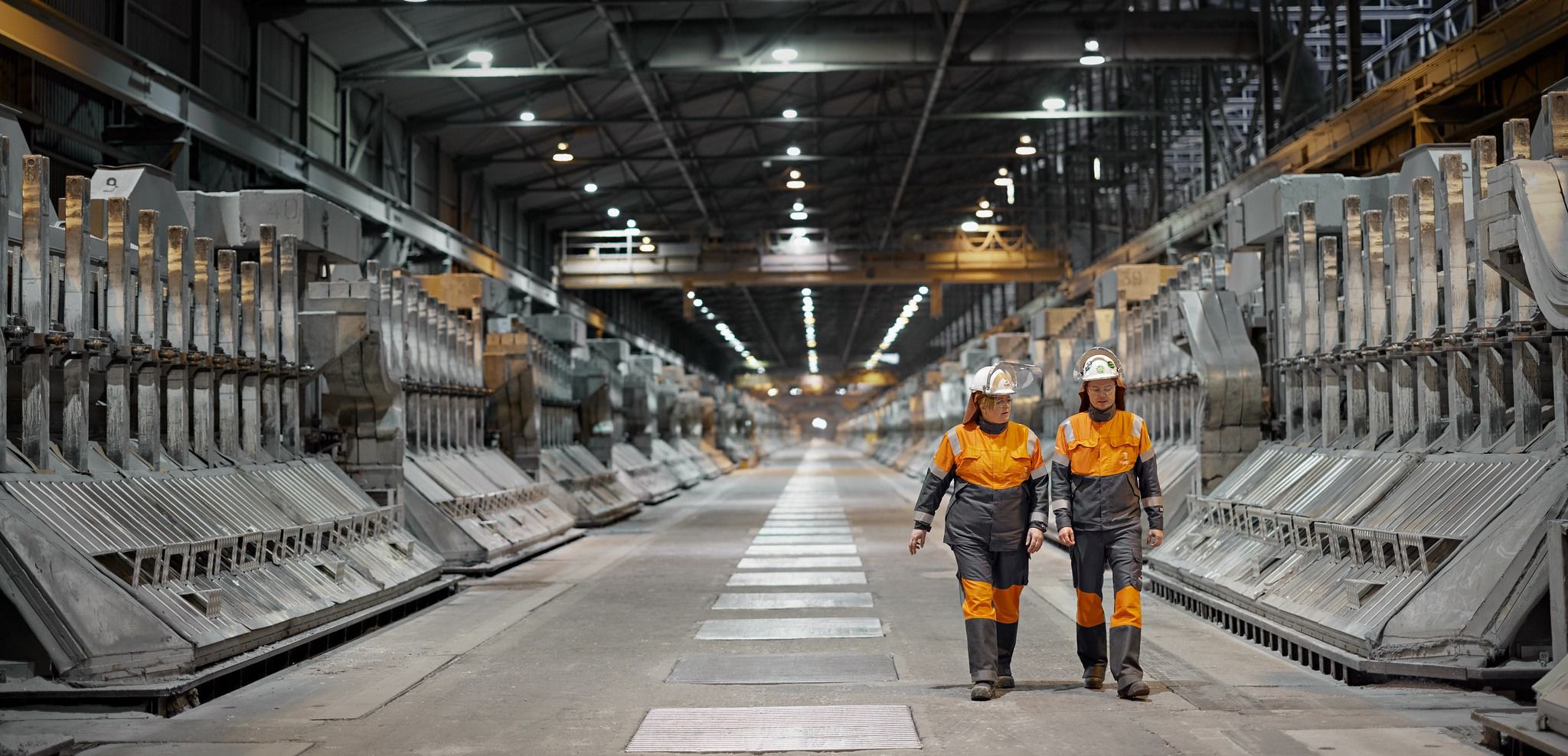 To arbeidere i oransje vernedrakter inne i en stor fabrikkhall