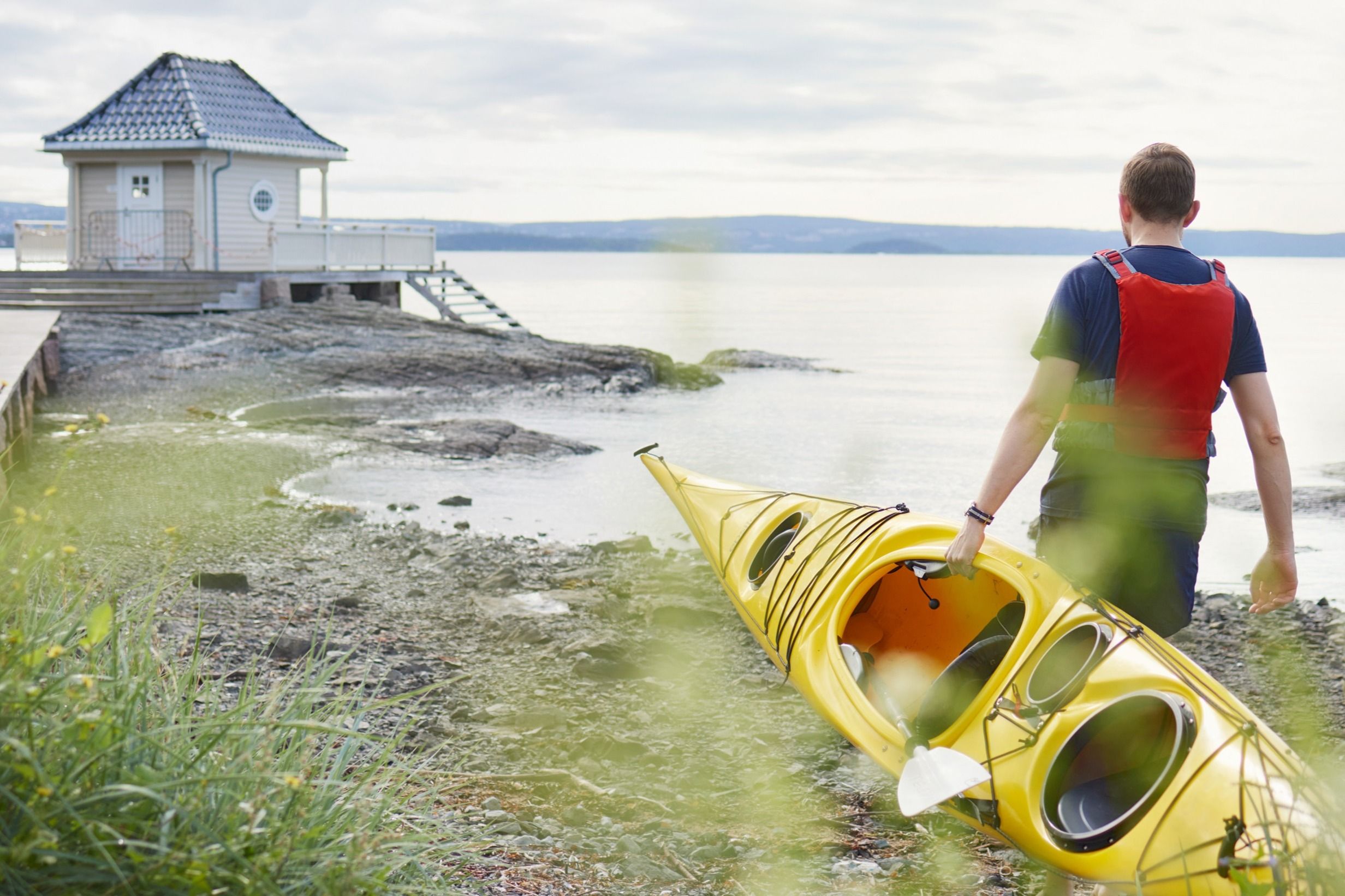 Mann bærer gul kajakk mot vannet ved et lite, hvitt badehus