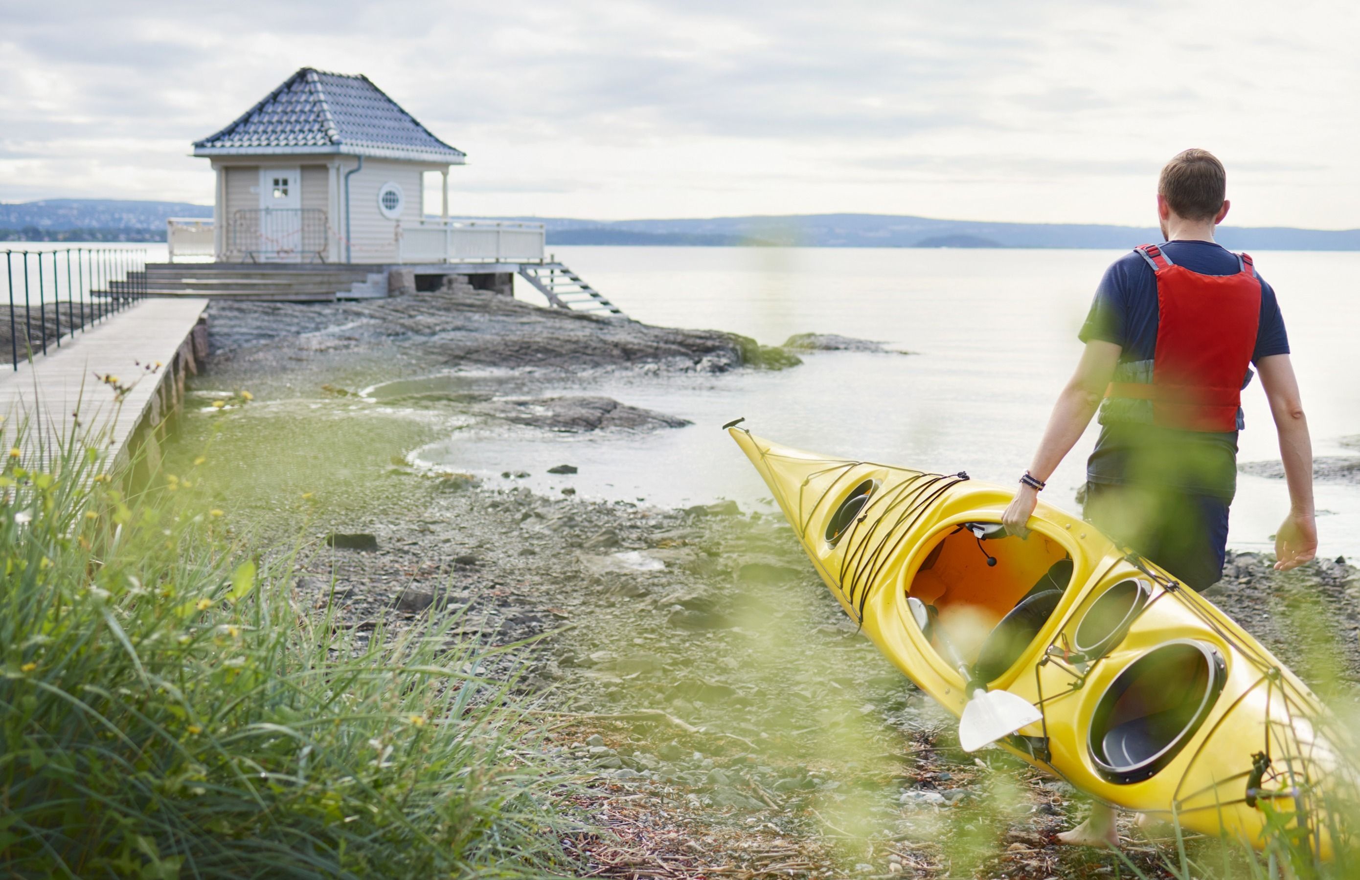 Mann bærer en gul kajakk langs steinete strand nær badehus