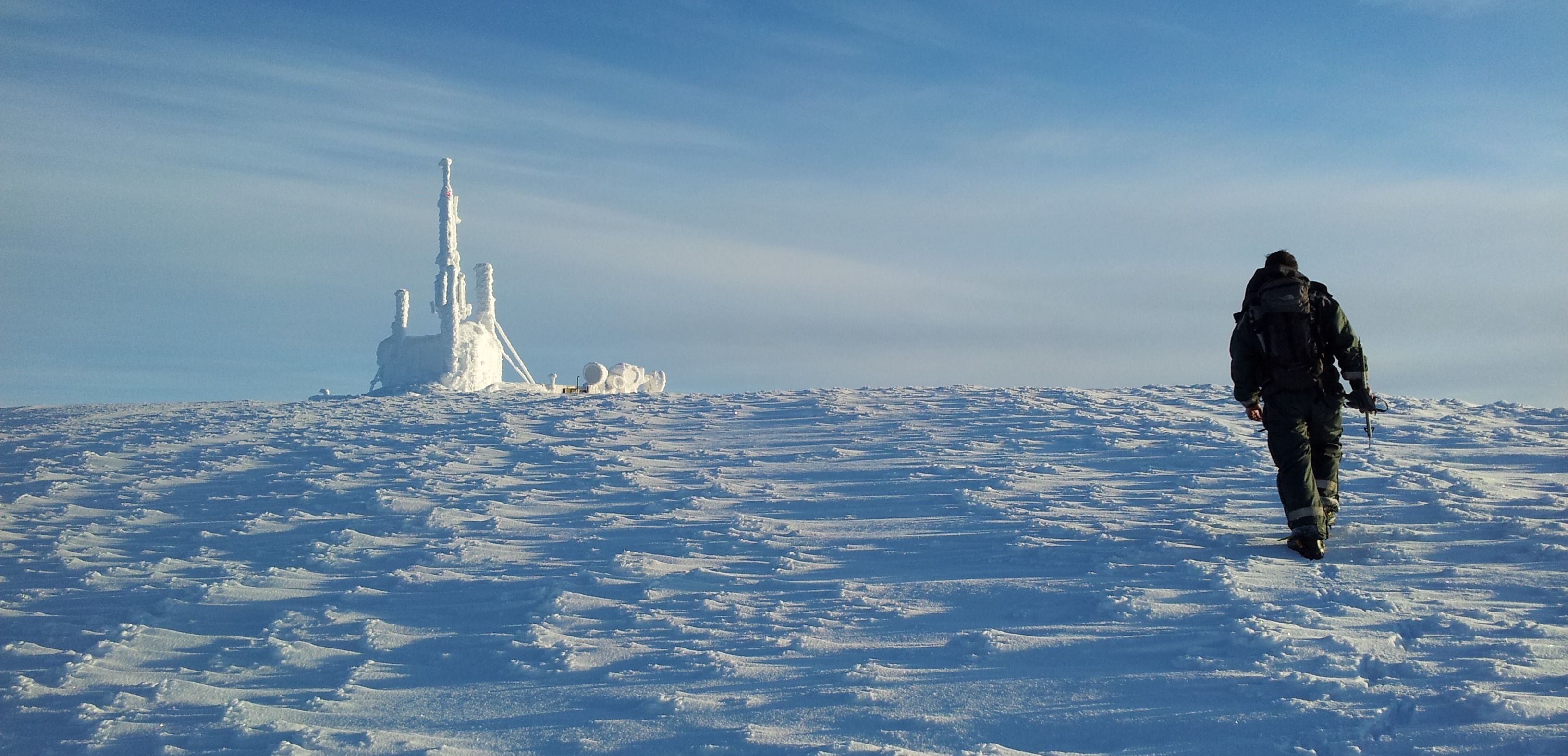 Person går mot isdekket struktur på snødekt slette under blå himmel