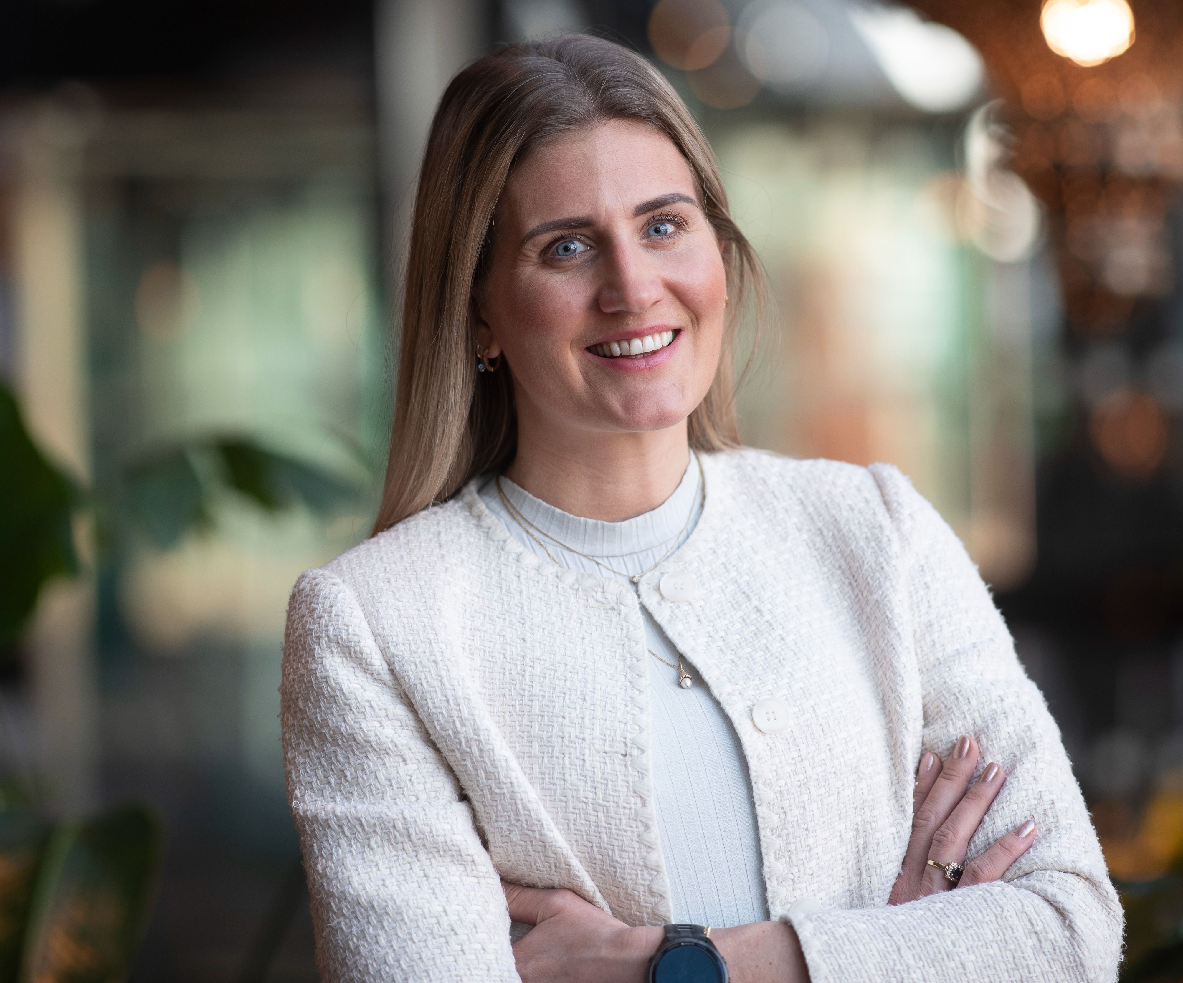 Person med langt blondt hår, iført hvit jakke og kors, står med armene i kryss