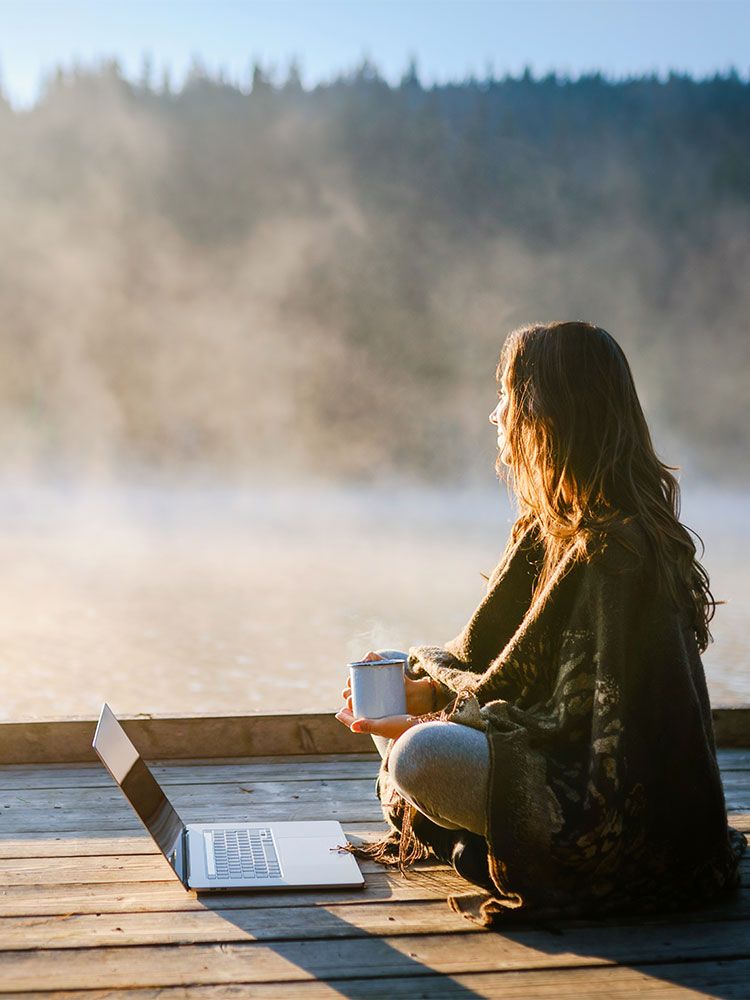 Kvinne med laptop og kaffekopp ved innsjø i morgendis