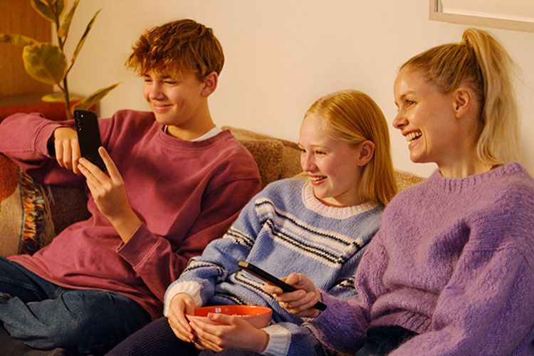 mor, datter og sønn sitter i sofaen og ser på tv med mobiltelefoner og en skål med popcorn