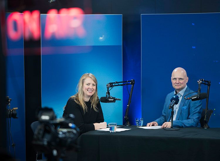To personer sitter i et radiostudio med mikrofoner og "ON AIR"-skilt