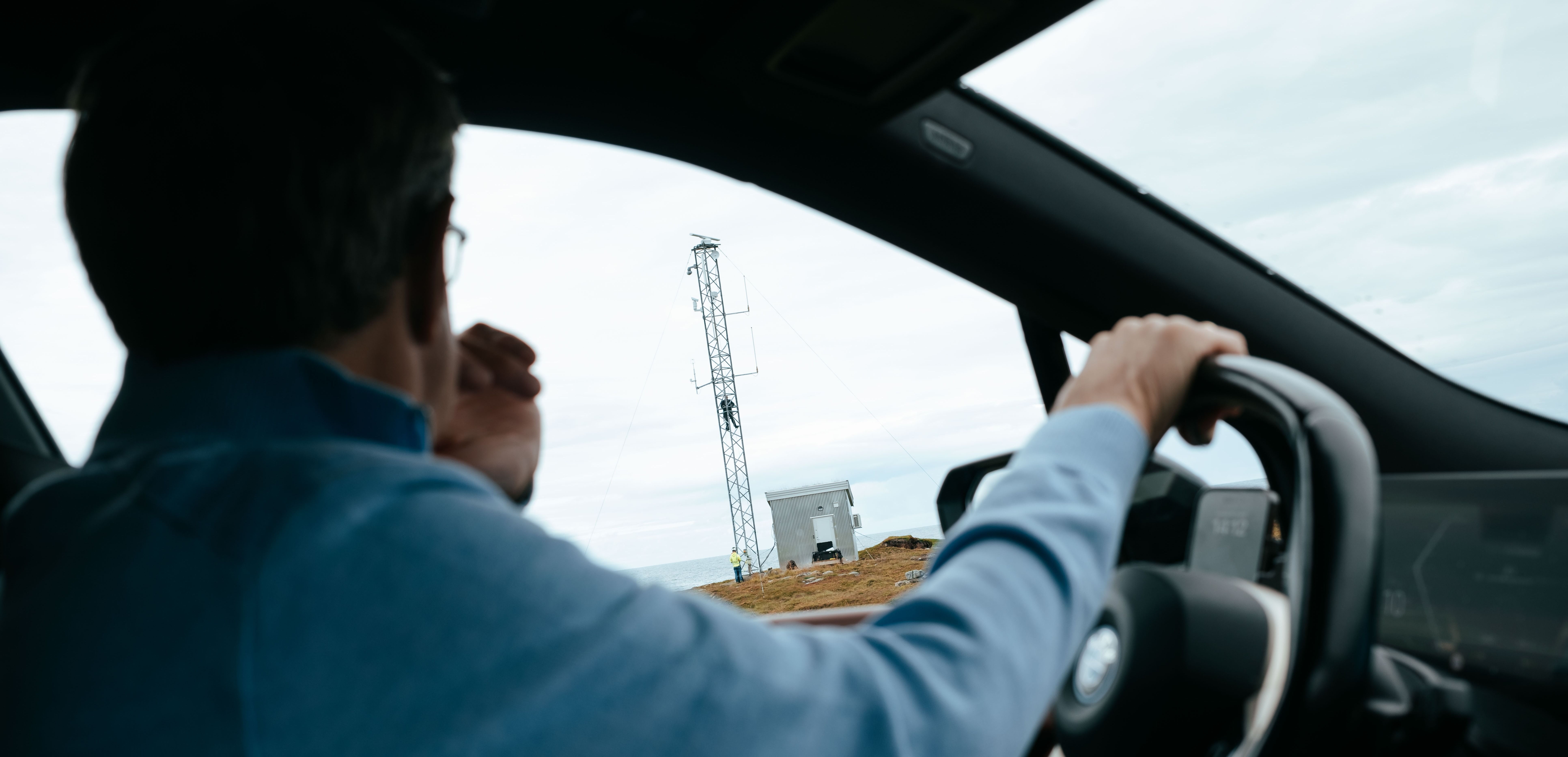 Mann kjører bil med utsikt til mast og lite bygg