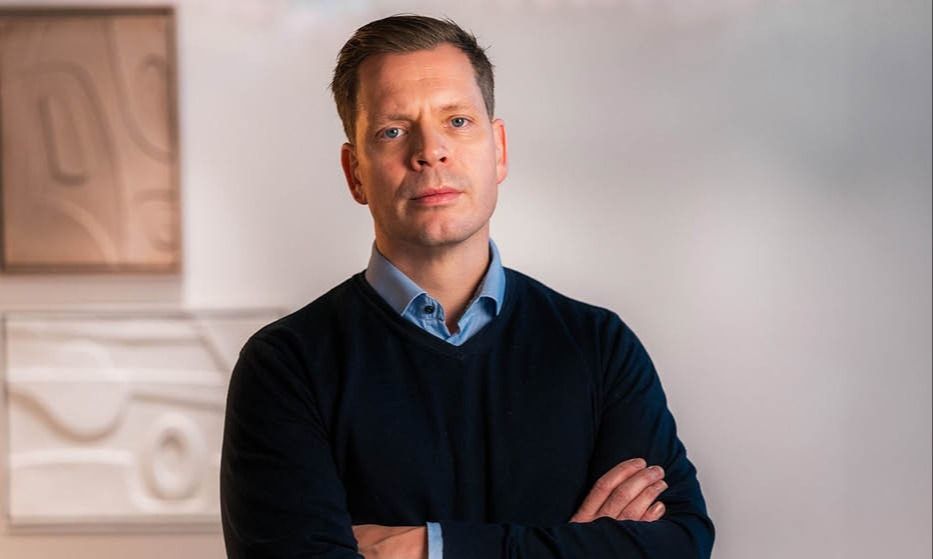 Person med blå skjorte og mørk genser, armene krysset i et lyst rom