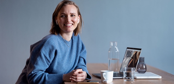 Smilende kvinne med ørepropper sitter ved et skrivebord med bærbar PC, mobil og kaffekopp.