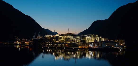 Opplyst industrikompleks ved vann om kvelden mellom fjell