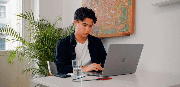 Person foran Macbook, plante og kunst på vegg